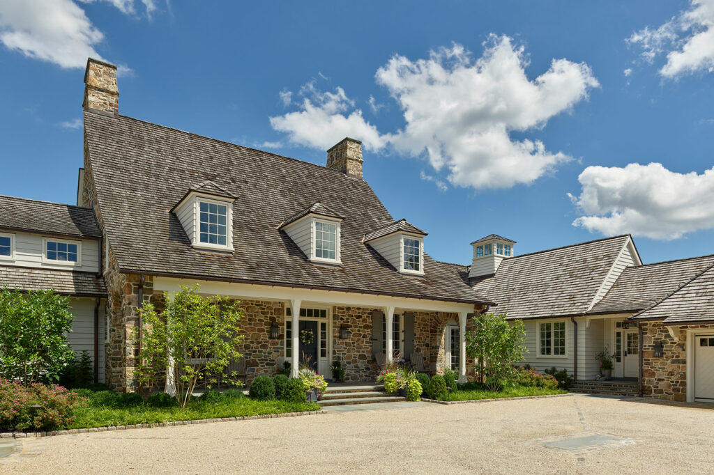 Entry court of cottage-style farmhouse with stone and siding exterior walls, front porch, sweeping cedar shake roof, dormers and cupola.