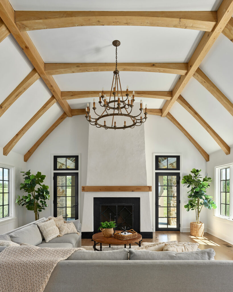 Family room in a contemporary farmhouse with white walls, black frame doors and windows, timber frame vaulted ceiling, and fireplace with wood mantel.